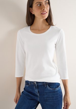 Weißes Langarmoberteil mit Rundhalsausschnitt, kombiniert mit dunkelblauen Slim-Fit-Jeans, die eine glatte Textur und einen lässigen Schnitt aufweisen.