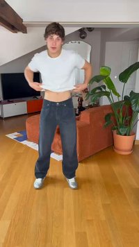 Čierne denimmové nohavice s voľným strihom, so zapínaním na gombík a zips, nosené s bielym tričkom. Drevená podlaha, zelené rastliny a oranžová pohovka v pozadí.