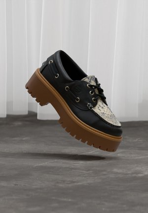 Zapato náutico de cuero negro con patrón de piel de serpiente beige y suela gruesa de tacos marrón flotando sobre suelo de hormigón con fondo de cortina blanca.