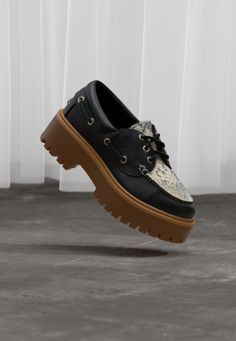 Chaussure bateau en cuir noir avec motif en peau de serpent beige et semelle épaisse à crampons marron, flottant au-dessus d'un sol en béton avec un fond de rideau blanc.