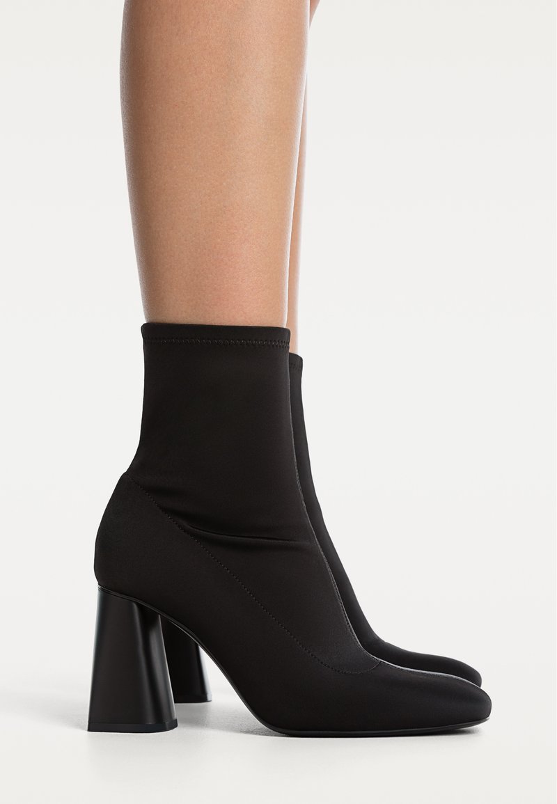 Černé ponožkové boty po lýtka s kulatou špičkou a silným vysokým podpatkem, obuté na holé nohy na bílém pozadí.