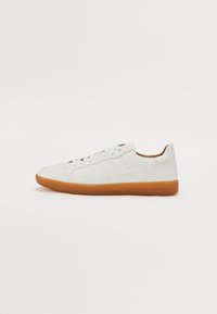 Sneaker en cuir blanc avec une texture lisse, bout arrondi, six oeillets et une semelle en caoutchouc beige. Présente des accents perforés sur les côtés.