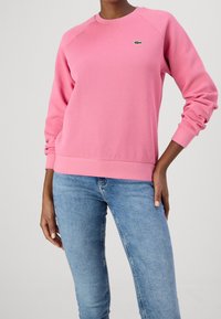 Różowa bluza z raglanowymi rękawami i zielonym logo. Posiada okrągły dekolt i teksturowaną tkaninę. Połączona z niebieskimi dżinsami.