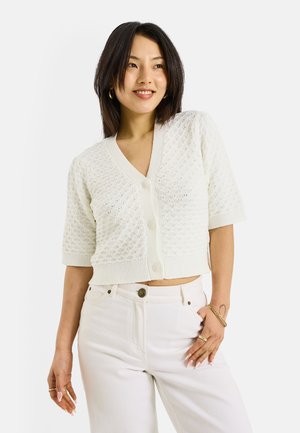 Mujer de cabello oscuro con una rebeca blanca texturizada de manga corta abotonada y pantalones blancos, sonriendo y posando con la mano en la cadera.