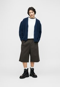 Νέος άνθρωπος στέκεται με τα χέρια στις τσέπες φορώντας ναυτικού μπλε ζακέτα, λευκό πουκάμισο, καφέ φαρδιά σορτς μέχρι τα γόνατα, λευκές κάλτσες και μαύρα μποτάκια.