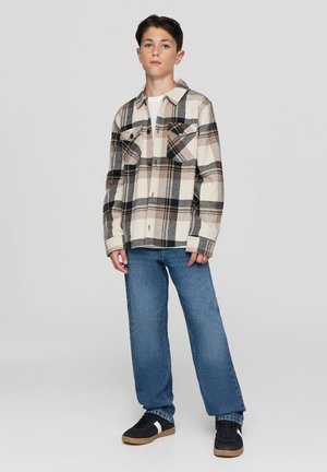 Camicia di flanella a quadri in beige, nero e marrone, con due tasche sul petto e chiusura a bottoni. Indossata con jeans blu a gamba dritta e sneakers nere.