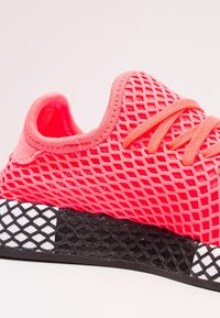 Ljusa rosa mesh-sneakers med texturerad ovandel, snörning och en svart-vit gummisula med rutmönster.