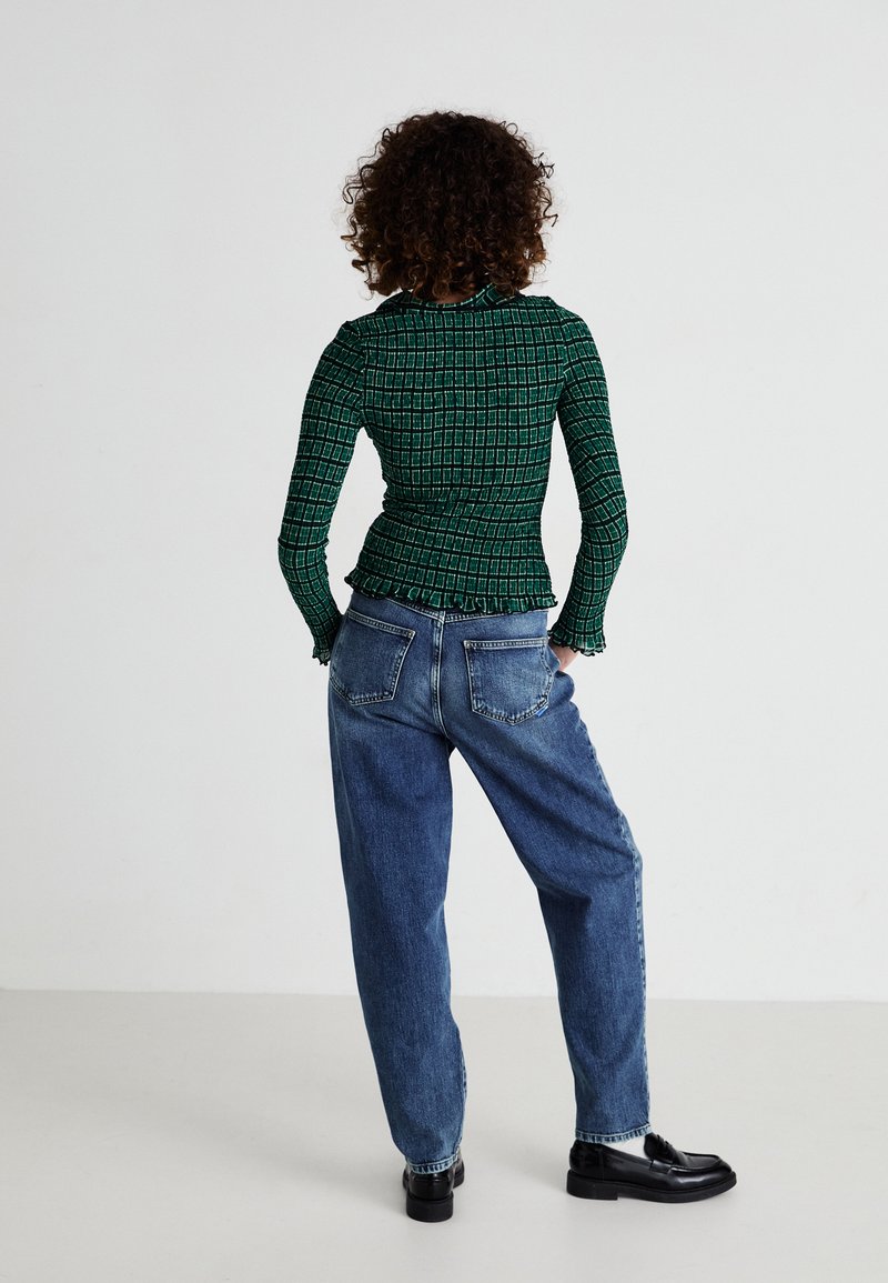 Grünes, gemustertes Langarmoberteil mit Rüschenrand, kombiniert mit hoch taillierten, lässig geschnittenen blauen Jeans und schwarzen Schuhen.