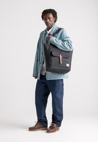 Crna torba s preklopom i kožnom trakom, napravljena od izdržljive tkanine, s prednjim džepom i oznakom logotipa, koju nosi osoba.