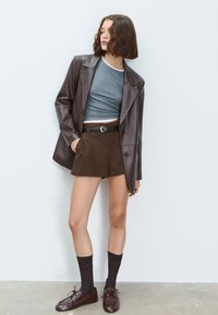 Blazer in finta pelle marrone sopra un top corto grigio, abbinato a pantaloni corti marroni, calze scure e scarpe marroni. Materiale morbido e vestibilità sartoriale.
