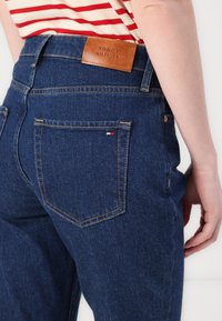Tamne plave traperice visokog struka, s stražnjim džepovima i smešnim kožnim oznakom TOMMY HILFIGER na pojasu.