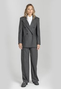 Szary dwurzędowy żakiet z dużymi klapami i czarnymi guzikami, w połączeniu z luźnymi szarymi spodniami i szarymi butami, na neutralnym tle.