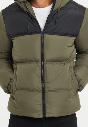 Man met een olijfgroene en zwarte gewatteerde jas met rits aan de voorkant en capuchon, handen in de zakken, over een wit T-shirt.