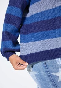 Gestrickter Pullover mit horizontalen Streifen in Dunkelblau, Hellblau und Grau, mit gerippten Bündchen und Saum, getragen über hellblauen Jeans.