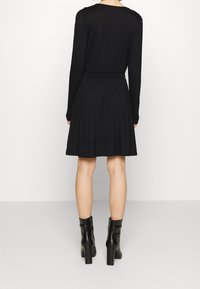 Femme portant une robe noire à manches longues arrivant aux genoux et des bottines noires à talons, vue de dos sur un fond uni.