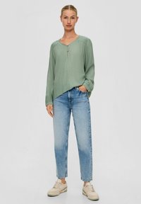 Lichtgroene, gestructureerde, lange mouw top met een V-hals en knopen, gecombineerd met middelhoge blauwe spijkerbroeken en beige sneakers.