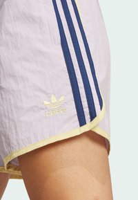 Helle lila Shorts mit gelbem Saum, drei dunkelblauen Streifen und einem gestickten goldenen Adidas-Logo auf der unteren linken Seite.