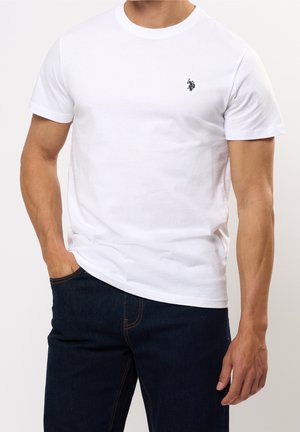 Mann trägt ein einfaches weißes T-Shirt mit kurzen Ärmeln und kleinem schwarzem Logo auf der Brust sowie dunkelblaue Jeans, Hand in der Tasche.