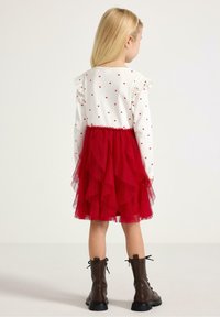 Roter, gestufter Tüllrock mit einem weißen Langarmoberteil, das kleine rote Herzmuster und Rüschenärmel hat. Braune Schnürstiefeletten.