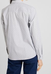 Camicia button-up grigia chiara con pois neri, maniche lunghe e dettagli sullo scollo posteriore. Il materiale sembra liscio con una sottile texture.