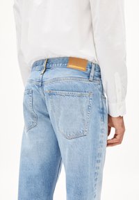 Jeans de mezclilla azul claro con un corte de tiro medio, bolsillos traseros y una etiqueta de cuero marrón con la inscripción "ARMEDANGELS" en la cinturilla.