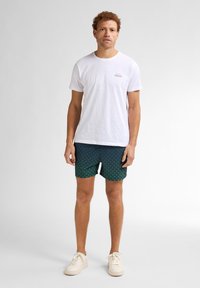 T-shirt bianca in cotone con un piccolo logo, abbinata a pantaloni da bagno a fantasie verdi e blu navy. Le scarpe sono sneakers di colore chiaro.