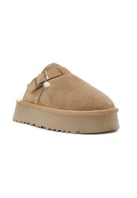 Tan suède slipper met een afgeronde teen, verstelbare gesp en witte bontvoering. Heeft een dikke platformzool met geribbelde textuur.