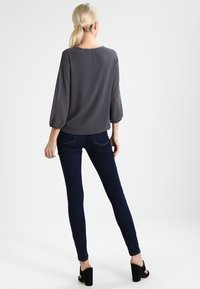 Graue Bluse mit dreiviertellangen Ballonärmeln, kombiniert mit dunklen, engen Jeans und schwarzen, hohen Sandalen. Glattes Material mit subtiler Satin-Optik.