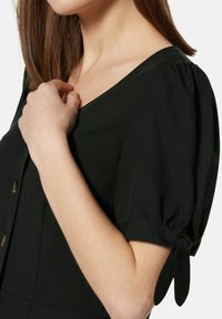 Schwarzes Kleid mit V-Ausschnitt, kurzen Puffärmeln mit Binddetail und goldenen Knöpfen entlang der Vorderseite. Glatte Textur, figurbetontes Design.