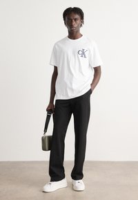 Hvid crew-neck t-shirt med blåt "CK" logo, parret med sorte bukser og hvide sneakers. Holder en lille grøn crossbody taske.