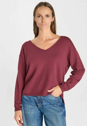 Donna con lunghi capelli castani che indossa un maglione bordeaux scollo a V morbido e jeans blu, con una mano in tasca, su uno sfondo semplice.