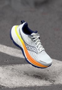 Baskets de course blanches avec une tige en mesh respirant, des accents noirs et jaunes, et une semelle rembourrée avec des détails orange et marine.