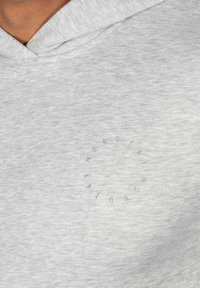 Sudadera gris claro hecha de material suave, con un bolsillo tipo canguro en la parte delantera y un logotipo gráfico circular en el pecho.