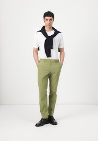 Camiseta blanca, pantalones verdes, suéter azul marino colgado sobre los hombros, zapatos negros, de pie frente a un fondo blanco liso.