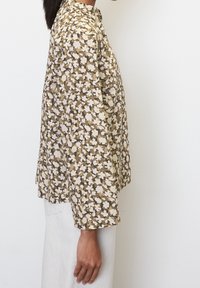 Blouse à motifs floraux dans des tons de marron et de crème, en tissu léger, à manches longues, avec une coupe décontractée, un col et des fentes sur les côtés.