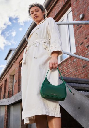 Hvid trenchcoat med knapper og en bundet talje, parret med en grøn læder skuldertaske med et logo og en buet form.