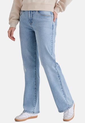 Person trägt hellblaue Jeans mit geradem Bein, beigefarbenen Sweatshirt und weiße Turnschuhe mit Kautschuksohlen, steht mit einer Hand in der Tasche.