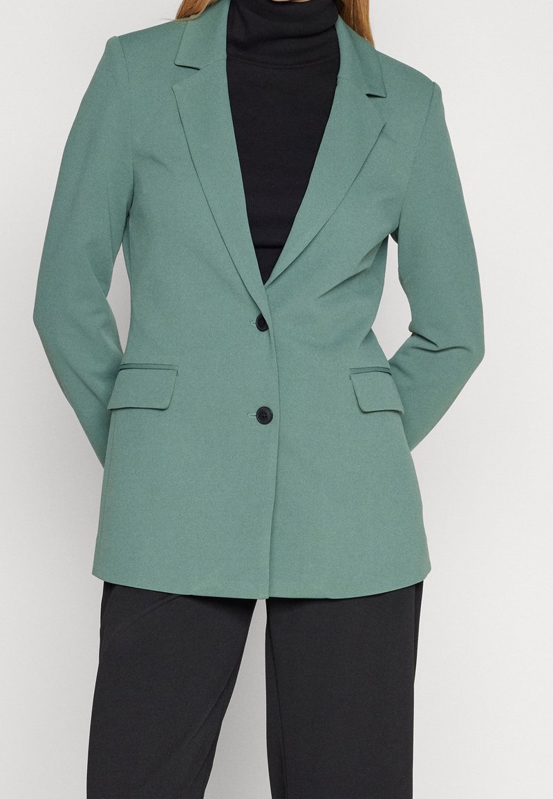 Femme portant un blazer vert cintré avec des boutons noirs sur un col roulé noir et un pantalon noir.