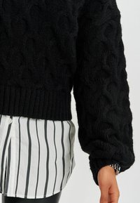Schwarzer gestrickter Pullover mit strukturiertem Zopfmuster, geripptem Saum und weiten Ärmeln, kombiniert mit einem gestreiften Hemd darunter.