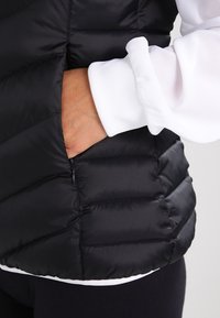 Černá prošívaná vesta s vodorovným prošíváním, s kapsou na zip a bílým manžetem vykukujícím ze spodní části.