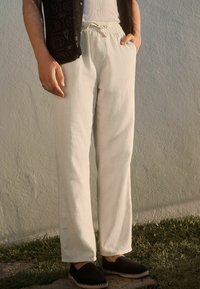 Pantalon beige clair à coupe décontractée, fabriqué à partir d'un tissu texturé, doté d'une taille élastique et d'un cordon de serrage. Associé à des chaussures noires à enfiler.