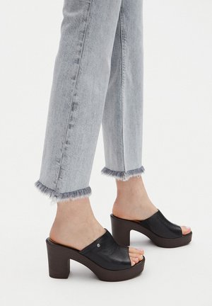 Schwarze Plateau-Mules aus Leder mit offenem Zehenbereich, einer abgerundeten Form und einem klobigen Absatz, der wie Holz aussieht. Gesehen zu ausgefransten grauen Jeans.