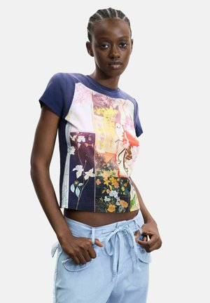 Kvinde med flettet hår iført en tætsiddende blomsterpatchwork crop top og lyseblå bukser med bindebånd i taljen, stående med hænderne nær lommerne.