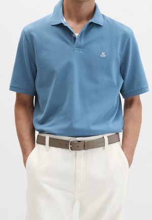 Homme portant un polo bleu clair à manches courtes avec le logo "MOP" et un pantalon blanc avec une ceinture beige, les mains dans les poches.