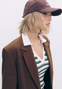 Brun blazer over en hvid og grøn stribet v-hals top. En brun kasket med et logo hviler på kort, bølget blond hår. Enkel baggrund.
