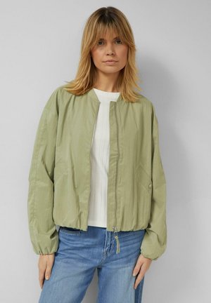 Kobieta o blond włosach sięgających ramion, ubrana w jasną zieloną kurtkę na zamek i biały top oraz niebieskie jeansy, stoi przy gładkiej ścianie.