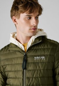 Chaqueta acolchada verde olivo con cierre de cremallera, que presenta un logo en el pecho, usada sobre una sudadera blanca y una camisa amarilla.