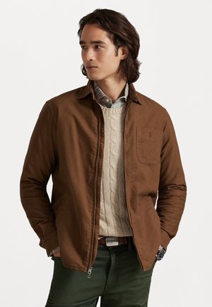 Chaqueta de algodón marrón con un cuello estructurado, bolsillo en el pecho y bolsillos laterales, superpuesta sobre un suéter de punto con trenzas color crema y una camisa a cuadros.