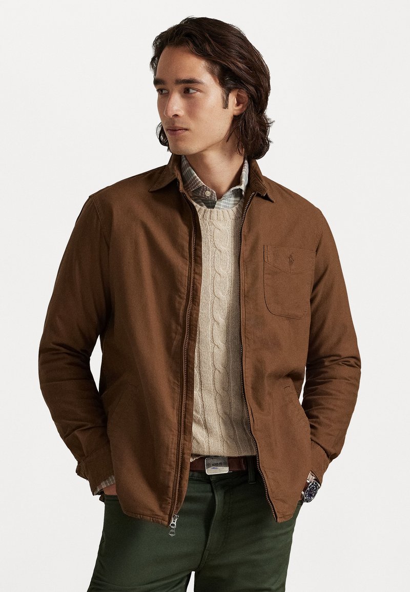 Chaqueta de algodón marrón con un cuello estructurado, bolsillo en el pecho y bolsillos laterales, superpuesta sobre un suéter de punto con trenzas color crema y una camisa a cuadros.