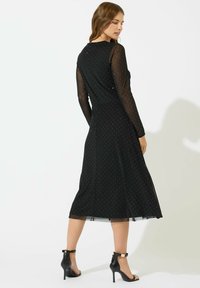 Abito nero con maniche lunghe in organza trasparente, motivo a pois, orlo svasato e corpetto attillato, abbinato a sandali neri con tacchi alti e cinturini.